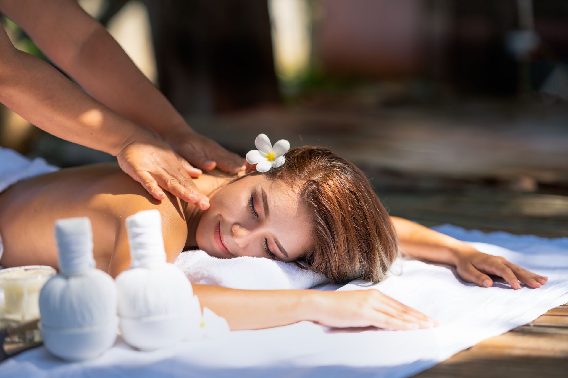 A woman receiving a massage outdoors, lying on a white towel, with a flower in her hair - Thames, NZ - Thai Classic Massage Thames