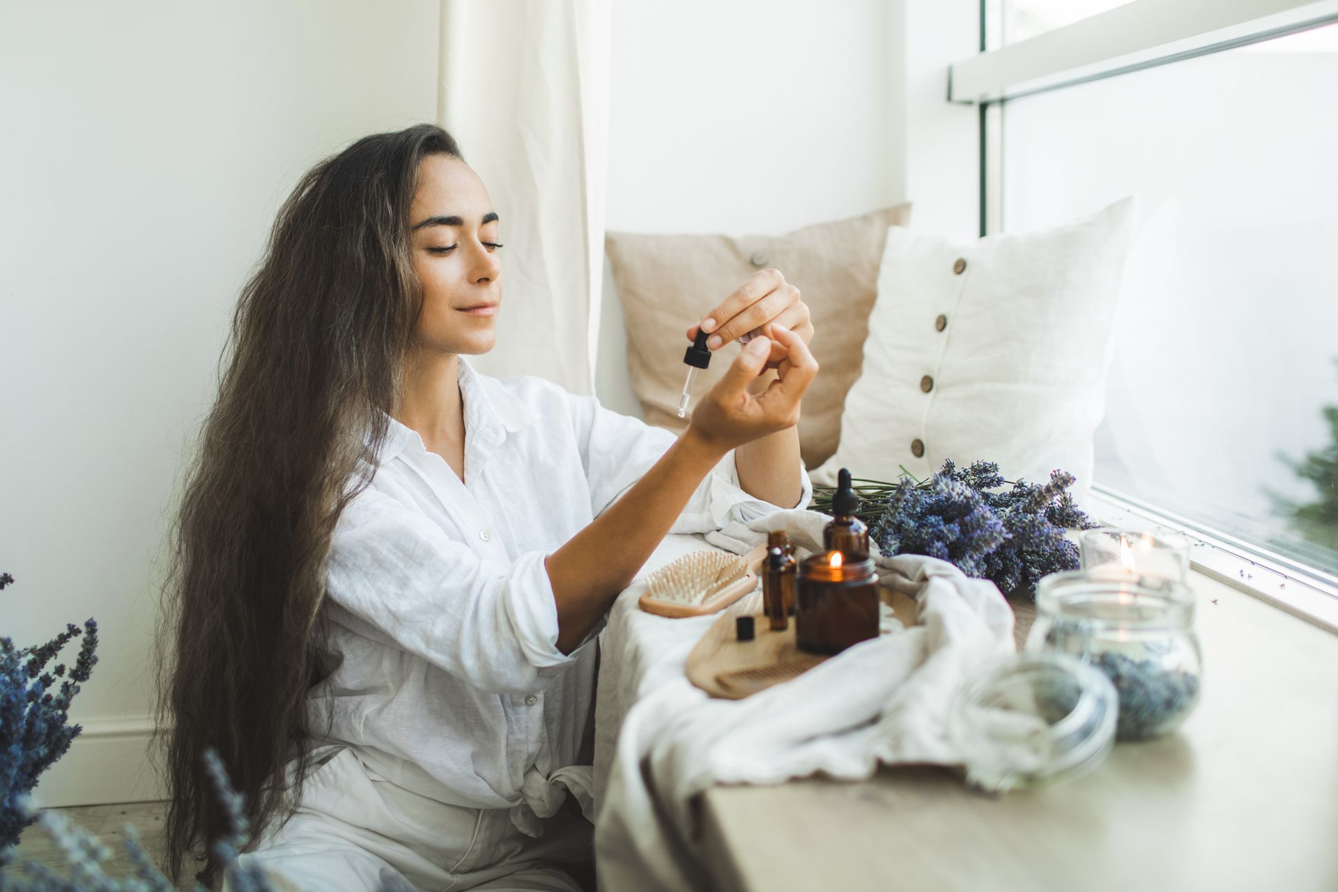A woman in a white robe uses a dropper bottle, surrounded by candles and lavender on a windowsill - Thames, NZ - Thai Classic Massage Thames