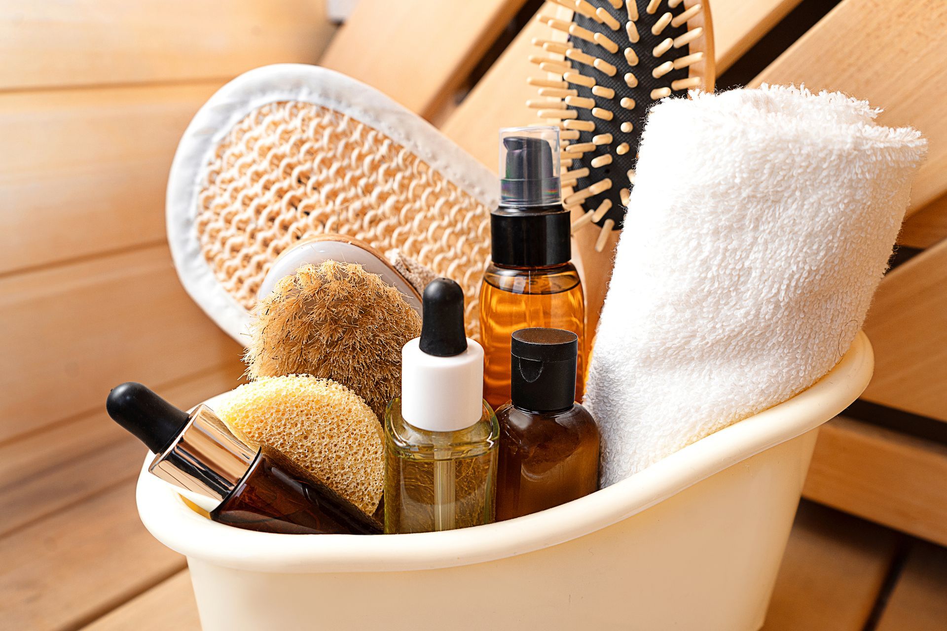 A white basin holding beauty products: brushes, loofahs, oils, and a rolled towel, against a wooden background - Thames, NZ - Thai Classic Massage Thames