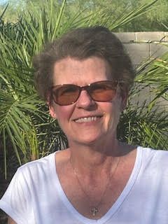 A woman wearing sunglasses and a white shirt is smiling in front of a palm tree.