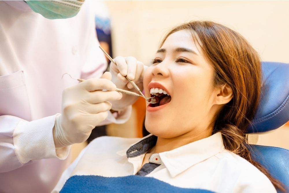 Woman in dentist chair with mouth open, being examined by dentist in white coat, gloves, and mask.
