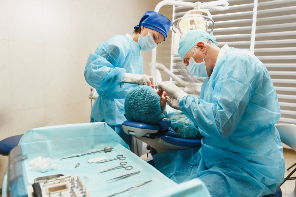 Dentists in blue scrubs and masks performing dental work on a patient in a dental chair, with surgical tools in view.