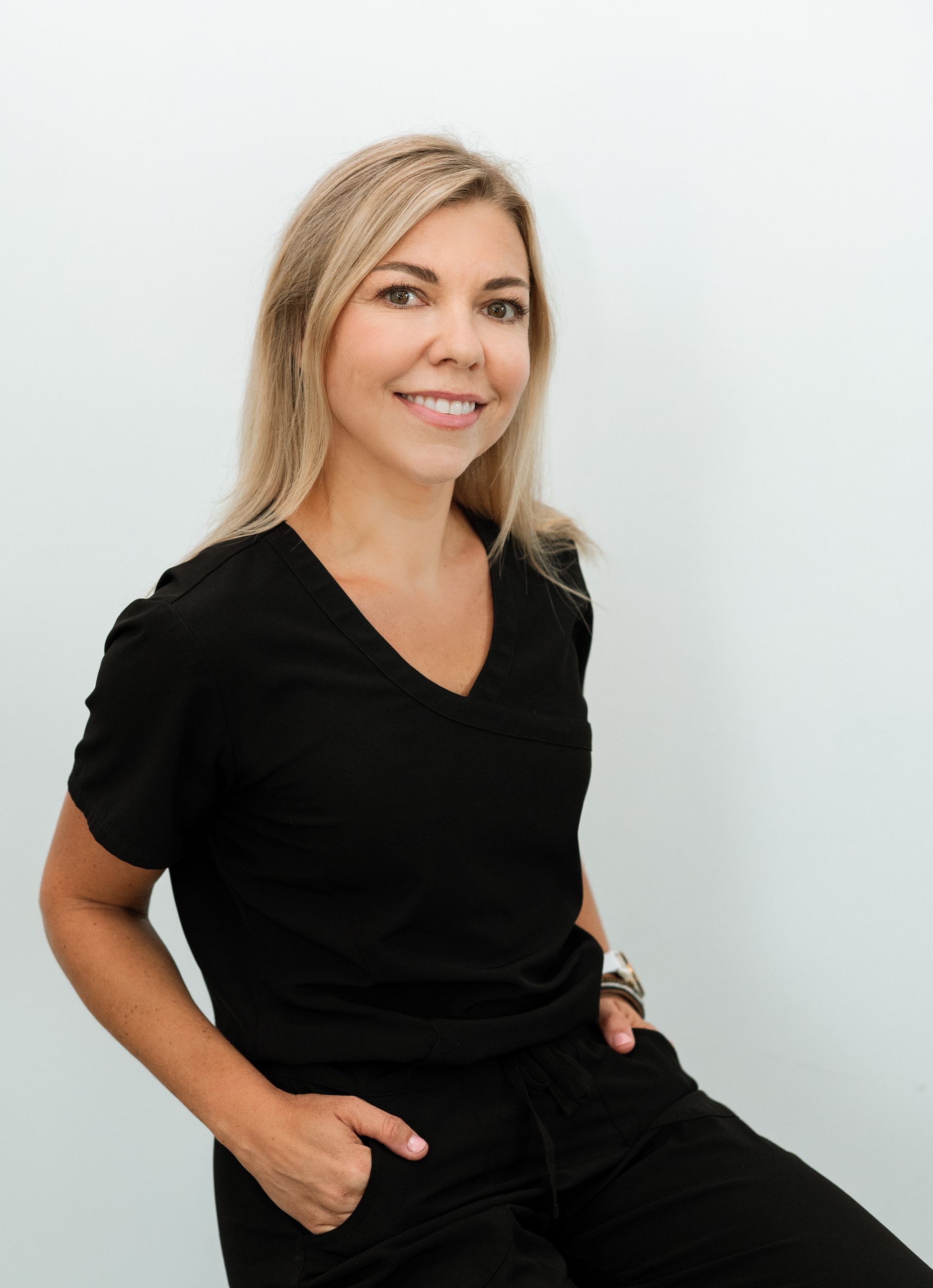 Woman with blonde hair in black scrubs, smiling, hands in pockets, leaning against a white wall.