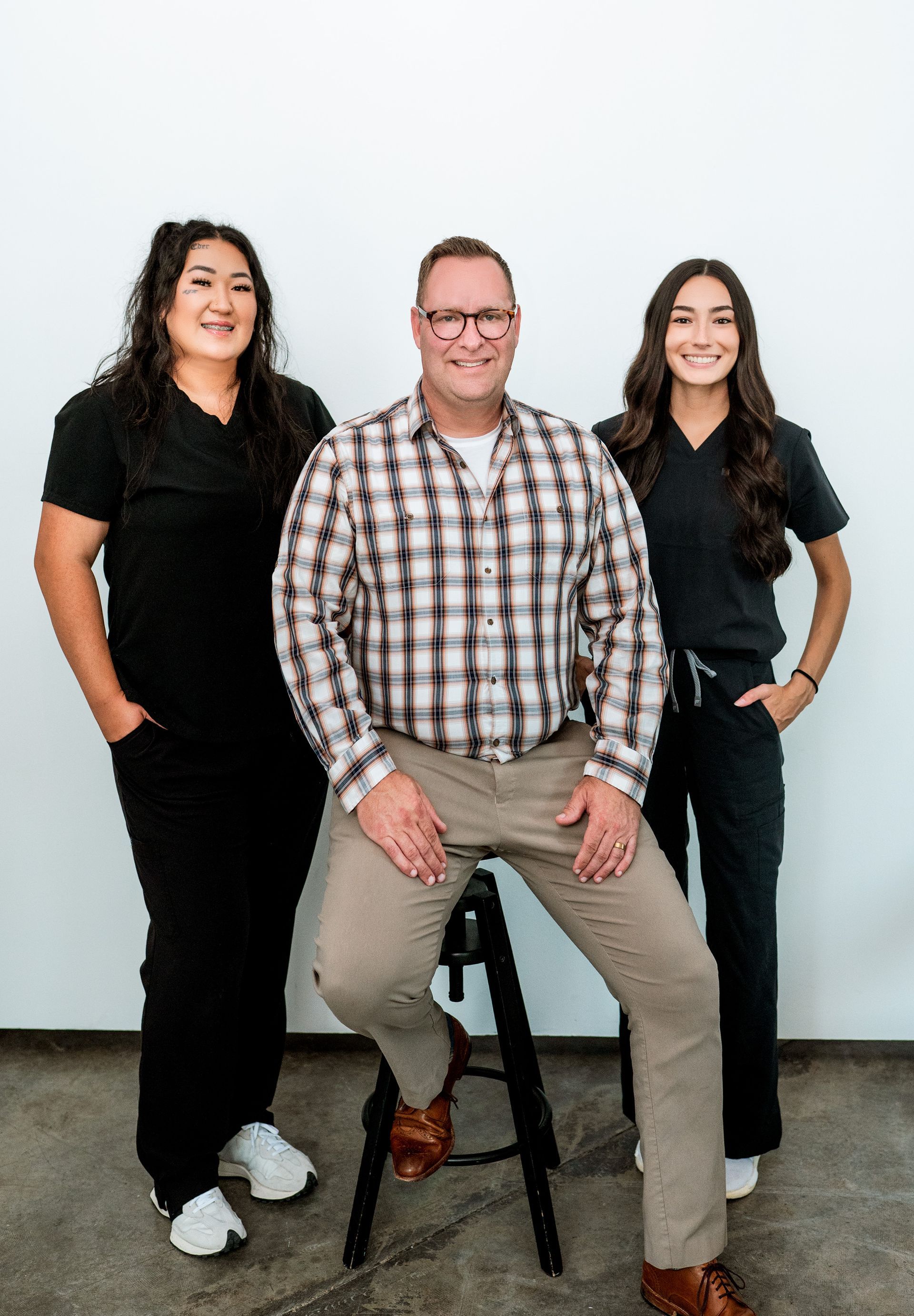 Three people: a man seated, wearing a plaid shirt; flanked by two women in black scrubs, all smiling in front of a white wall.
