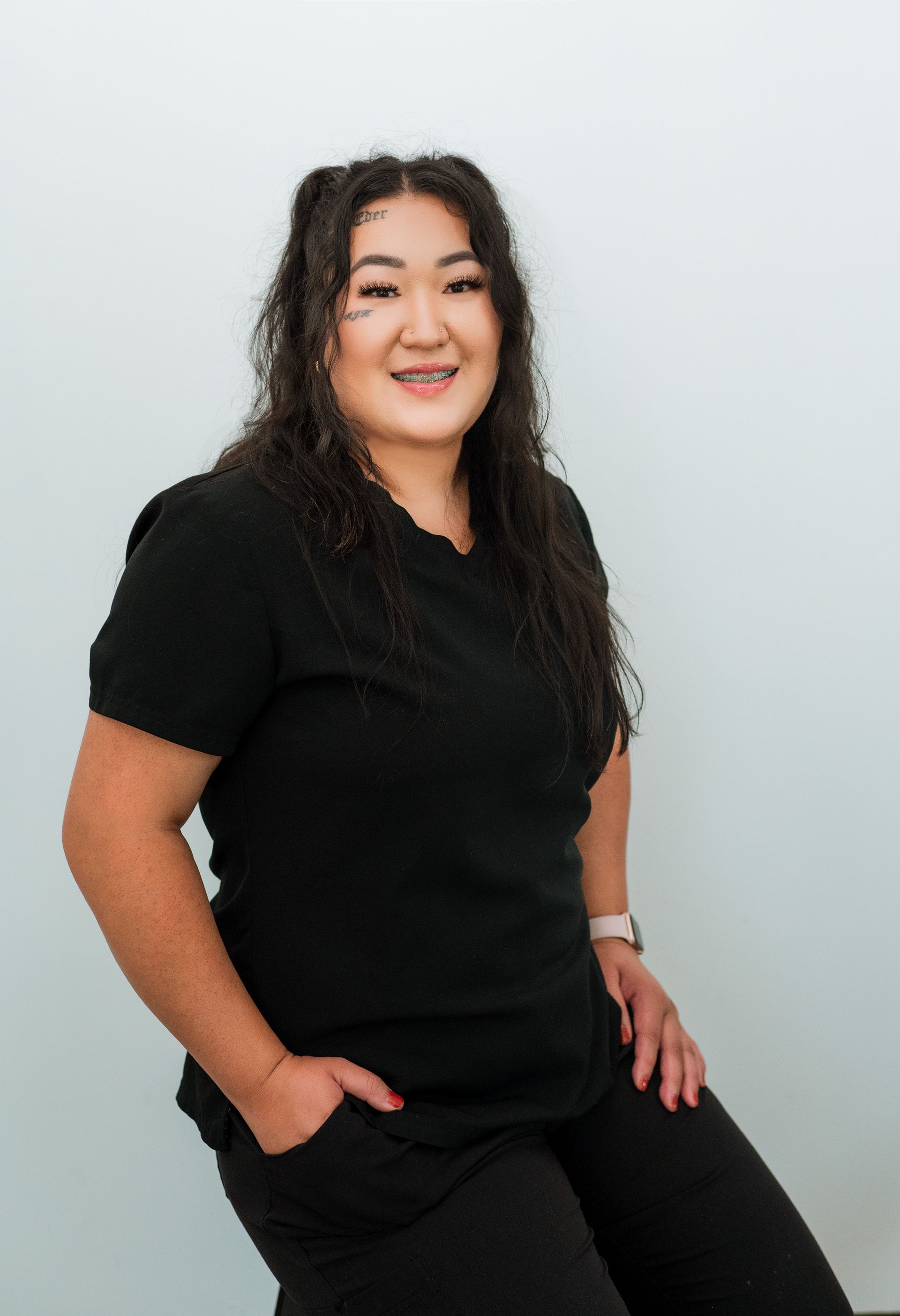 Woman with long dark hair in black scrubs, smiling, hands in pocket. Against a white background.