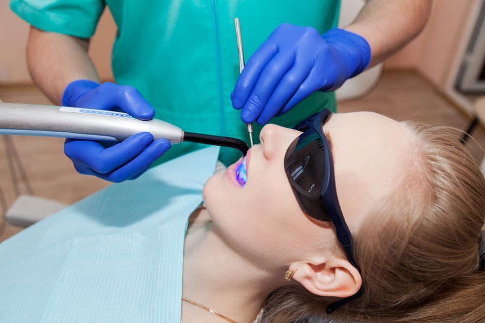 Dentist using a curing light on a patient's teeth; patient wearing safety glasses in a dental office.