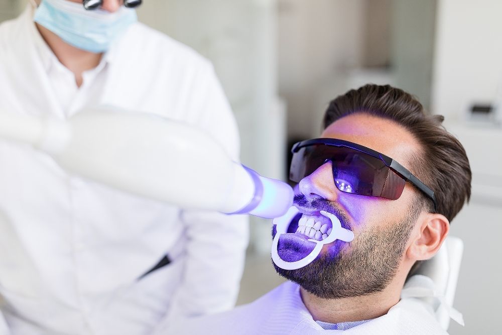 Man getting teeth whitened at a dental clinic, wearing protective glasses, being treated with a blue light.
