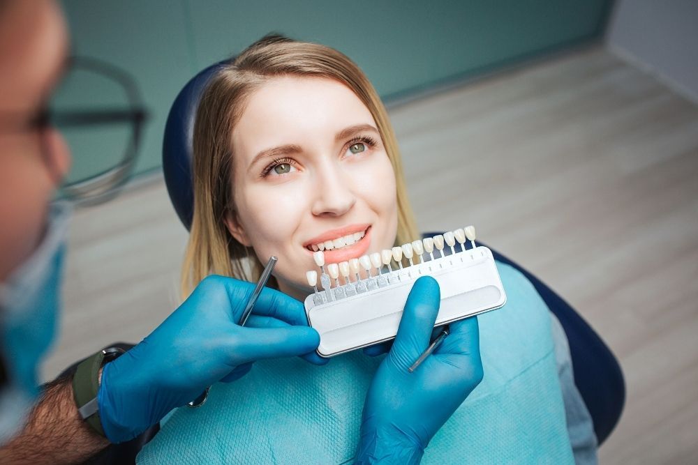 Woman at dentist's choosing teeth shade. Dentist holds shade guide, wearing gloves, in blue chair.