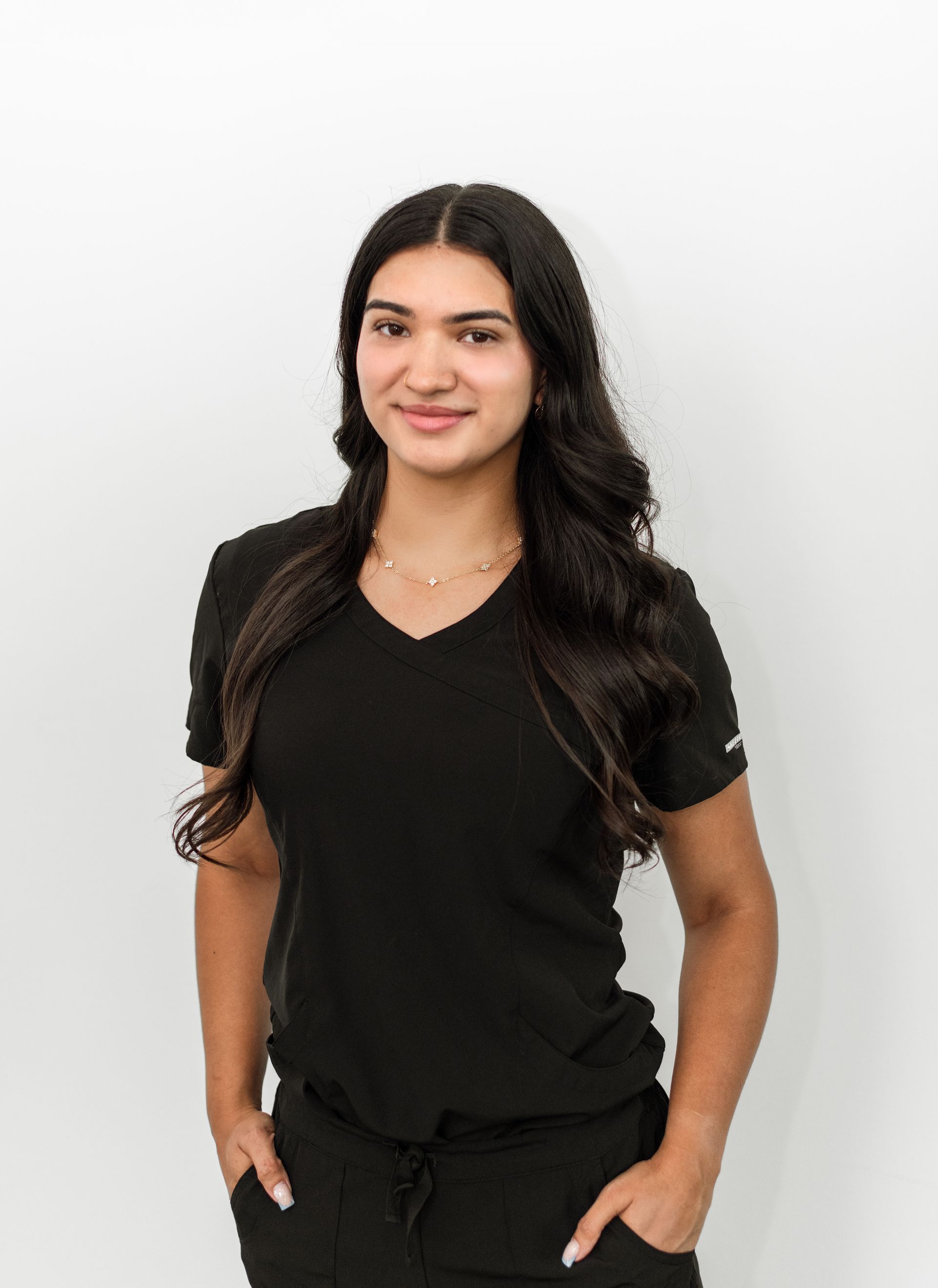 Young woman in black scrubs, hands in pockets, smiling against a white backdrop.
