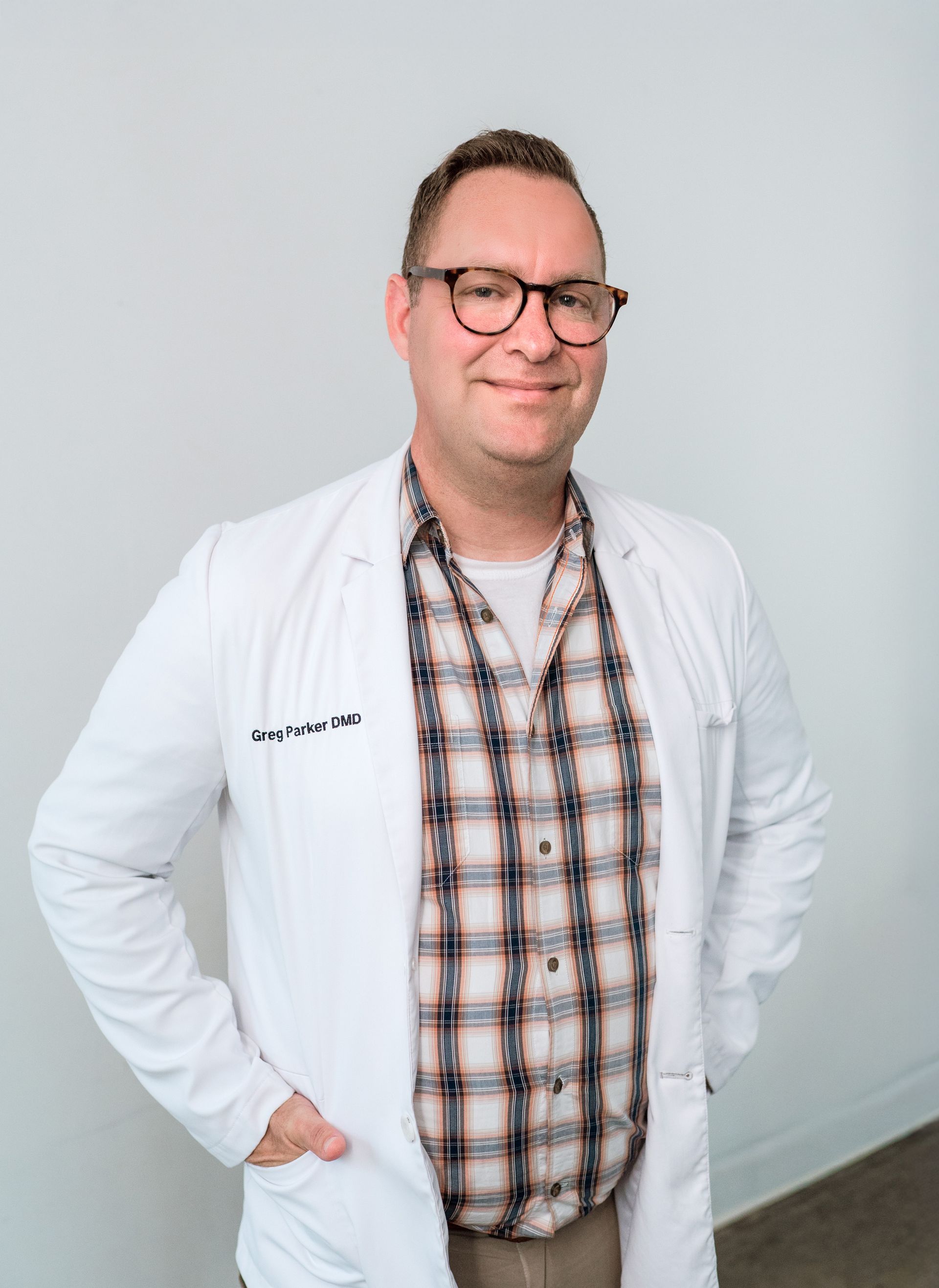 Man in glasses, white coat, plaid shirt, hands in pockets, smiling against white backdrop.