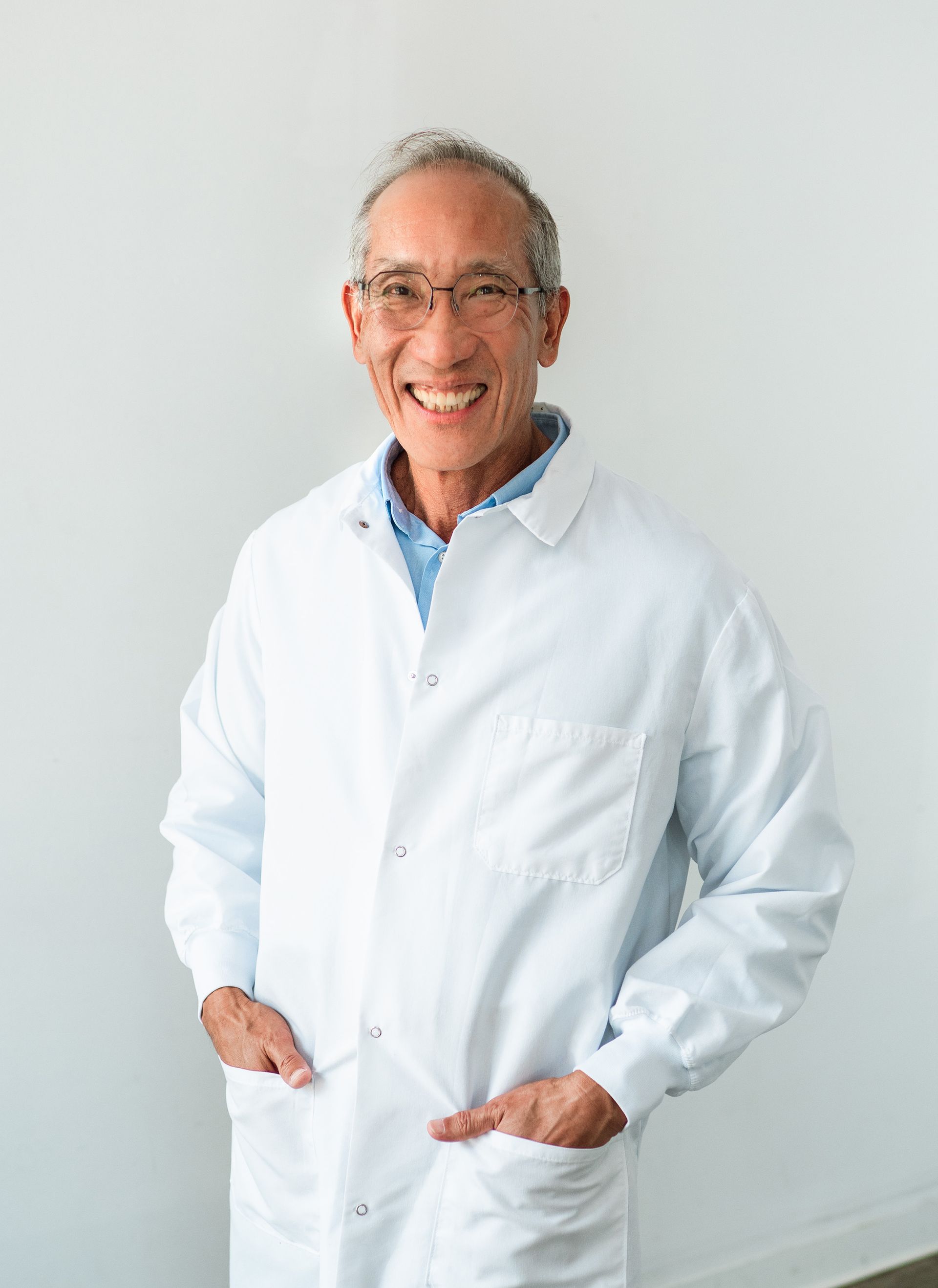 Man in white lab coat, smiling, leaning against white wall, hands in pockets.
