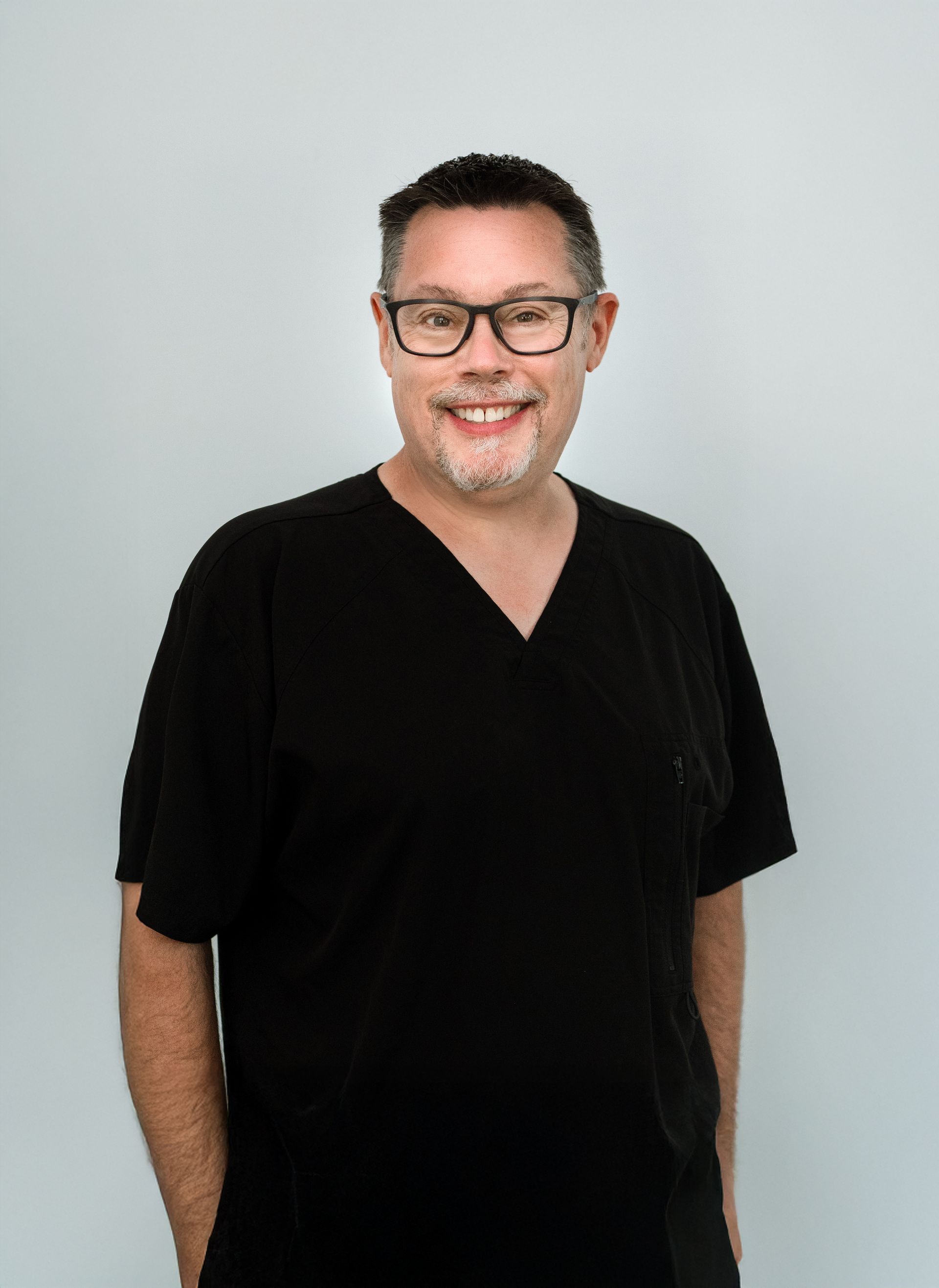 Man in black scrubs and glasses smiles at the camera, set against a white wall.