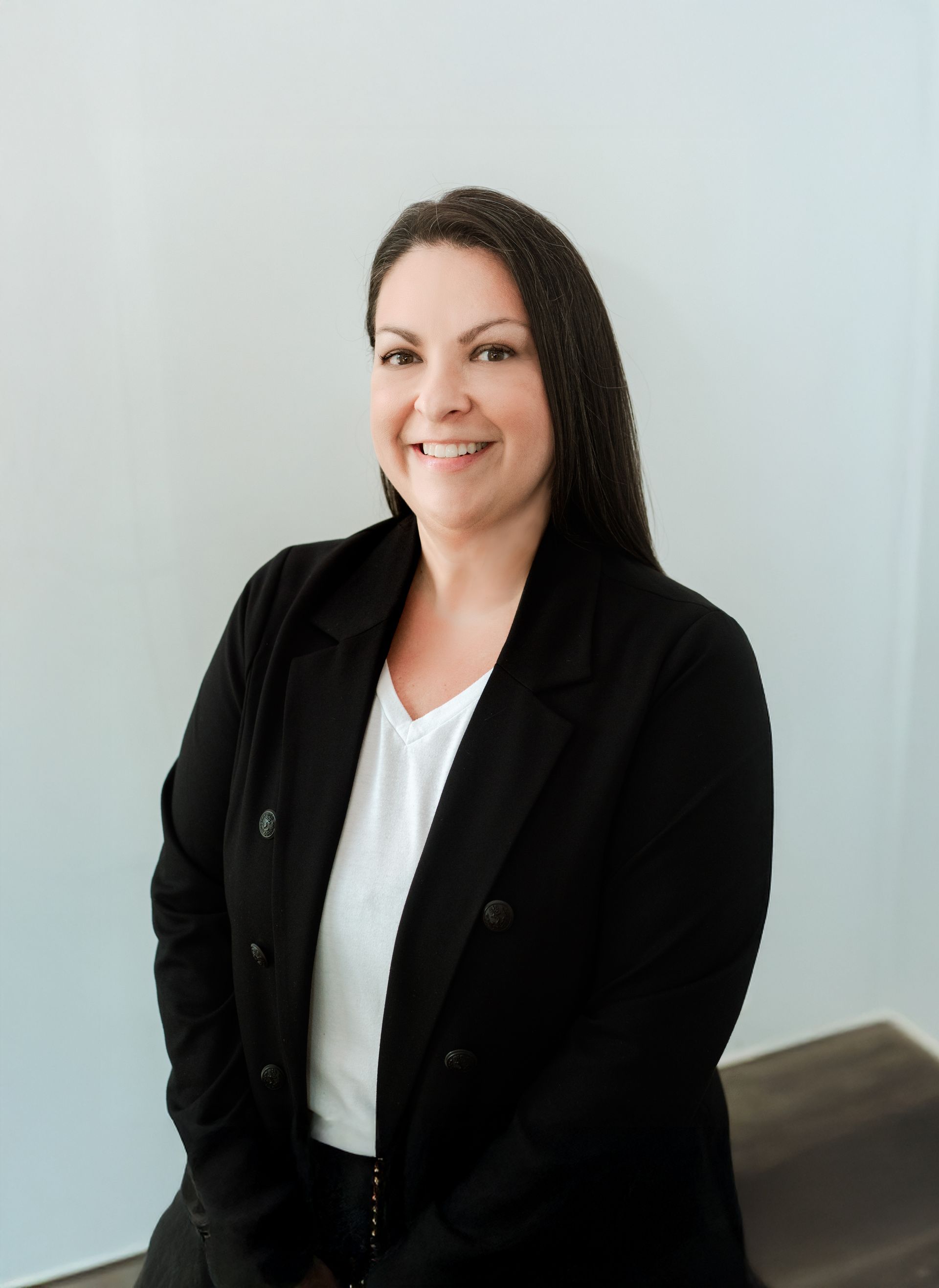 Woman with long dark hair, smiling, wearing a black blazer and white shirt, against a light-colored wall.
