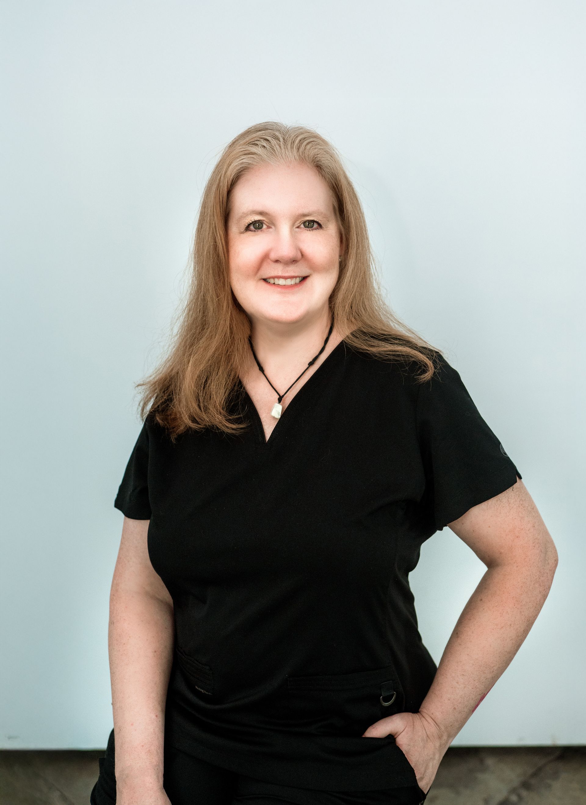 Woman with blonde hair, in black top, smiling, hand in pocket, posing in front of a white wall.