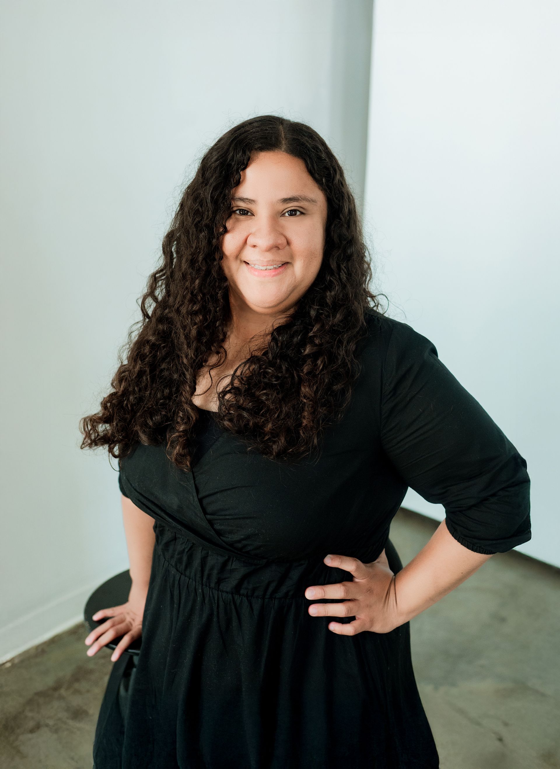 Woman with curly hair, smiling, wearing black dress, posing, hand on hip, leaning against white wall.
