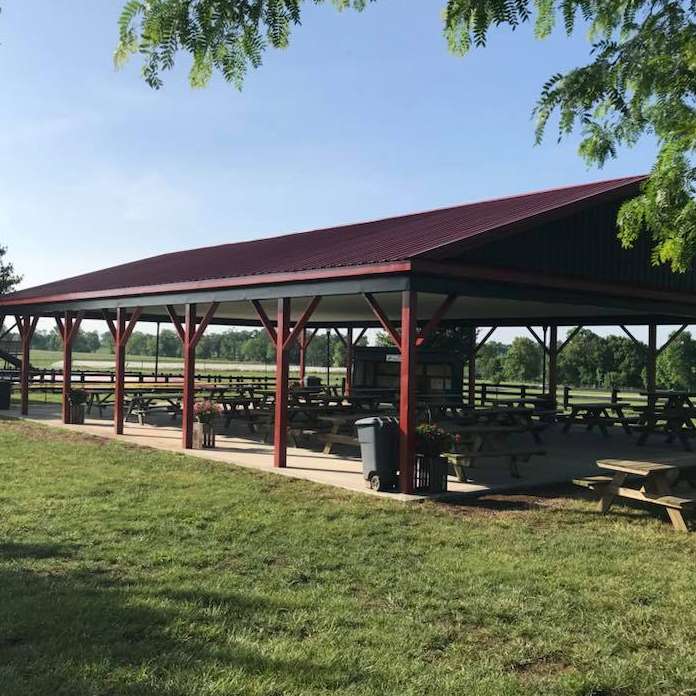 A large open-air pavilion with a burgundy roof and picnic tables, located in a grassy park.