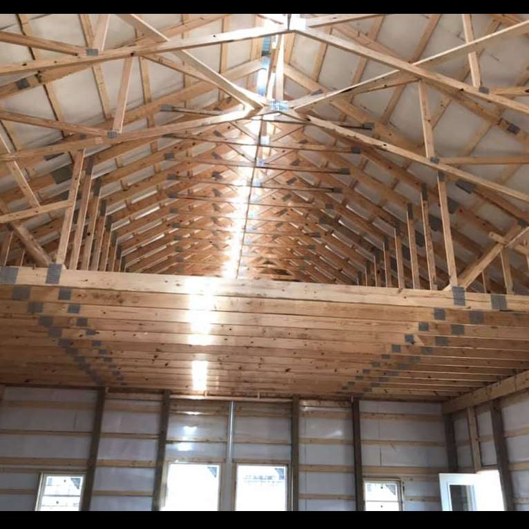 Inside view of a building under construction with a wooden truss roof. Sunlight shines through the peak of the roof onto the wooden beams and floor.