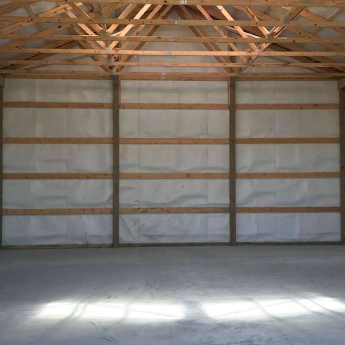 Interior view of a large, empty building with a concrete floor and wooden framing. The walls are insulated with white material.