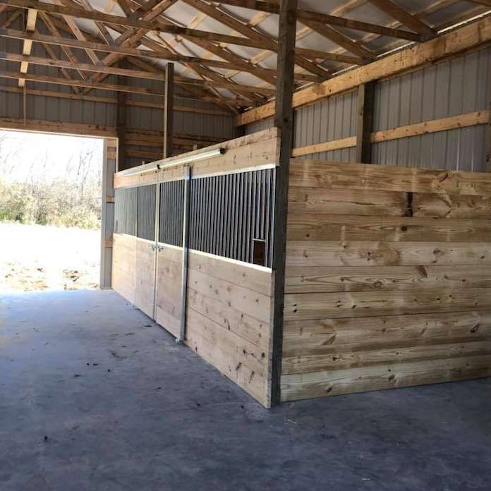 Inside a barn, wooden horse stalls with metal bars and sliding doors are visible. The barn has a concrete floor and a wooden roof.