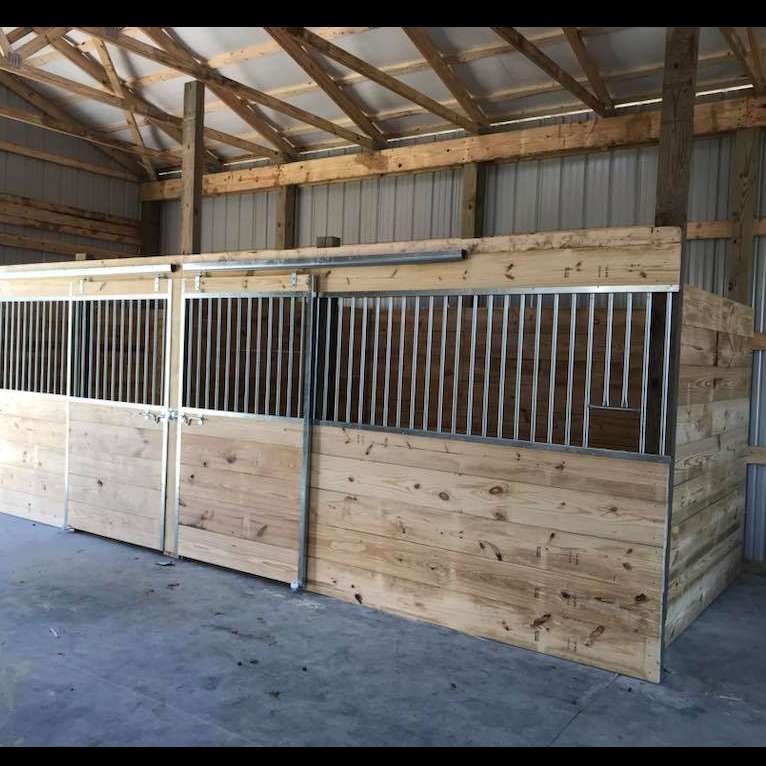Horse stalls inside a wooden barn with metal bars and sliding doors. The stalls are light-colored wood against a gray concrete floor.