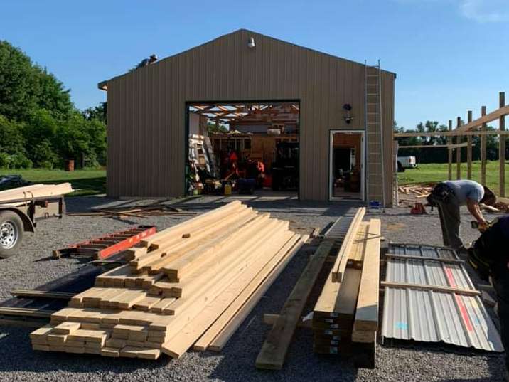 Construction of a tan metal building. Lumber and materials are in the foreground, workers are visible, with an open garage door and side door.