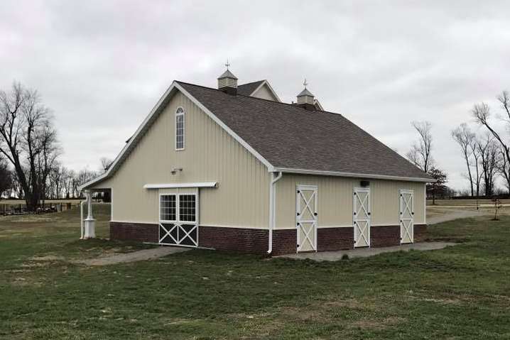 Tan barn with a brown roof, white trim, and three white doors. It is set on a green lawn under a cloudy sky.
