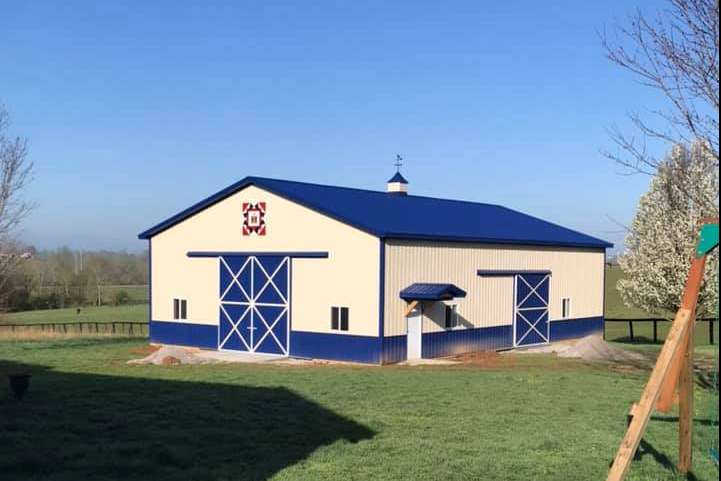 A cream-colored barn with a blue roof and trim, standing in a grassy yard under a clear blue sky.