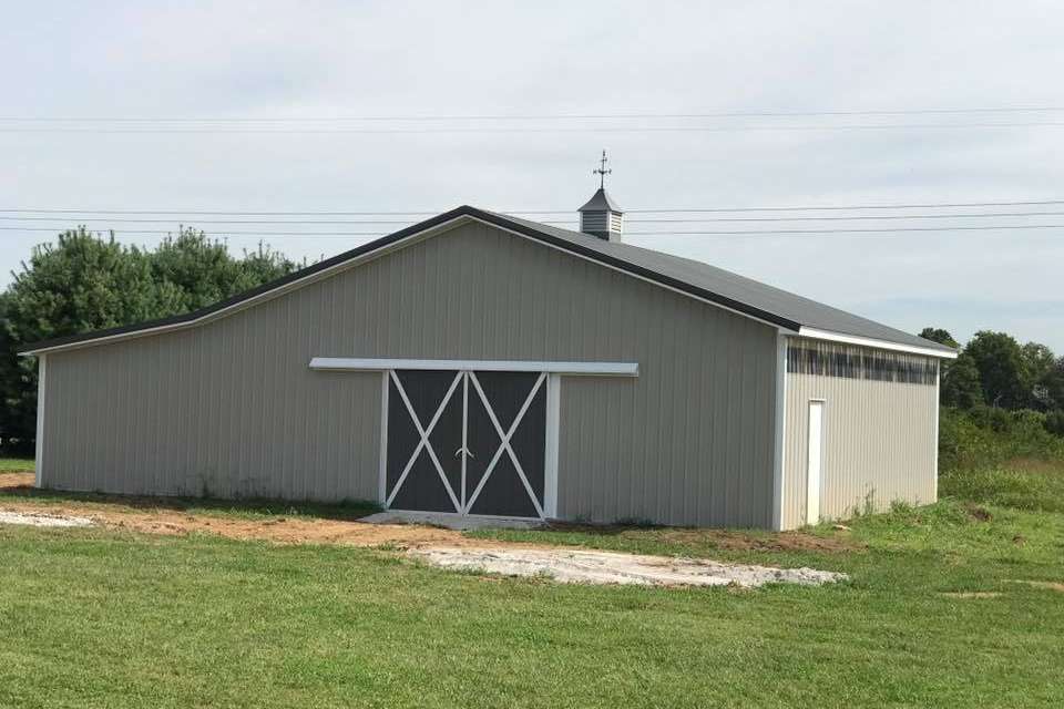 Gray barn with a black roof and sliding doors, located in a grassy field under a blue sky.