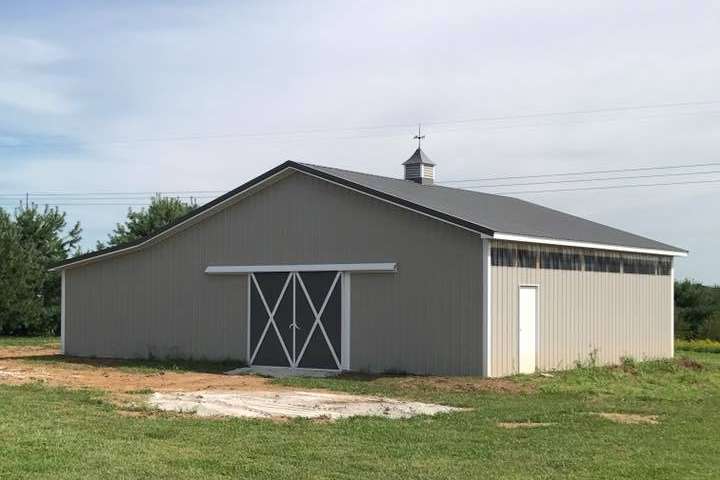A gray barn with a dark roof sits on a grassy field. Two sliding doors with white trim and a small door are on the front.