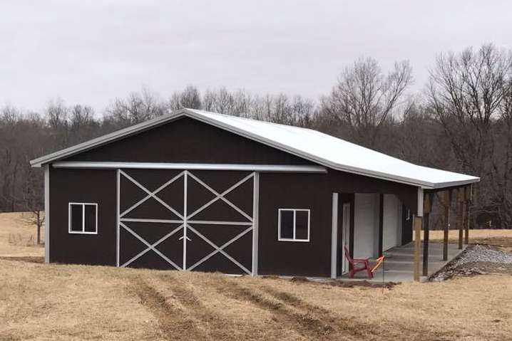 Dark brown barn with a white roof and sliding doors, and a small porch. It sits in a field with trees in the background.