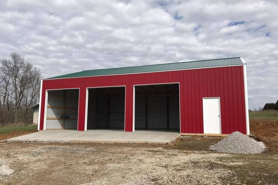 Red metal garage with three bays and a white door, set on a concrete pad under a cloudy sky.