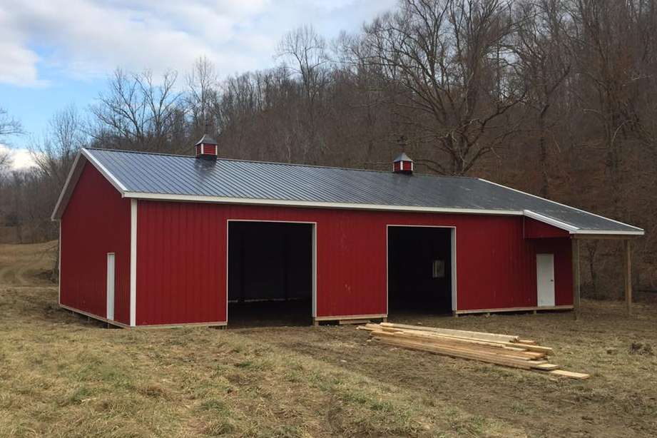Red barn with two large openings and a smaller covered area. The barn has a gray metal roof and is set in a field.