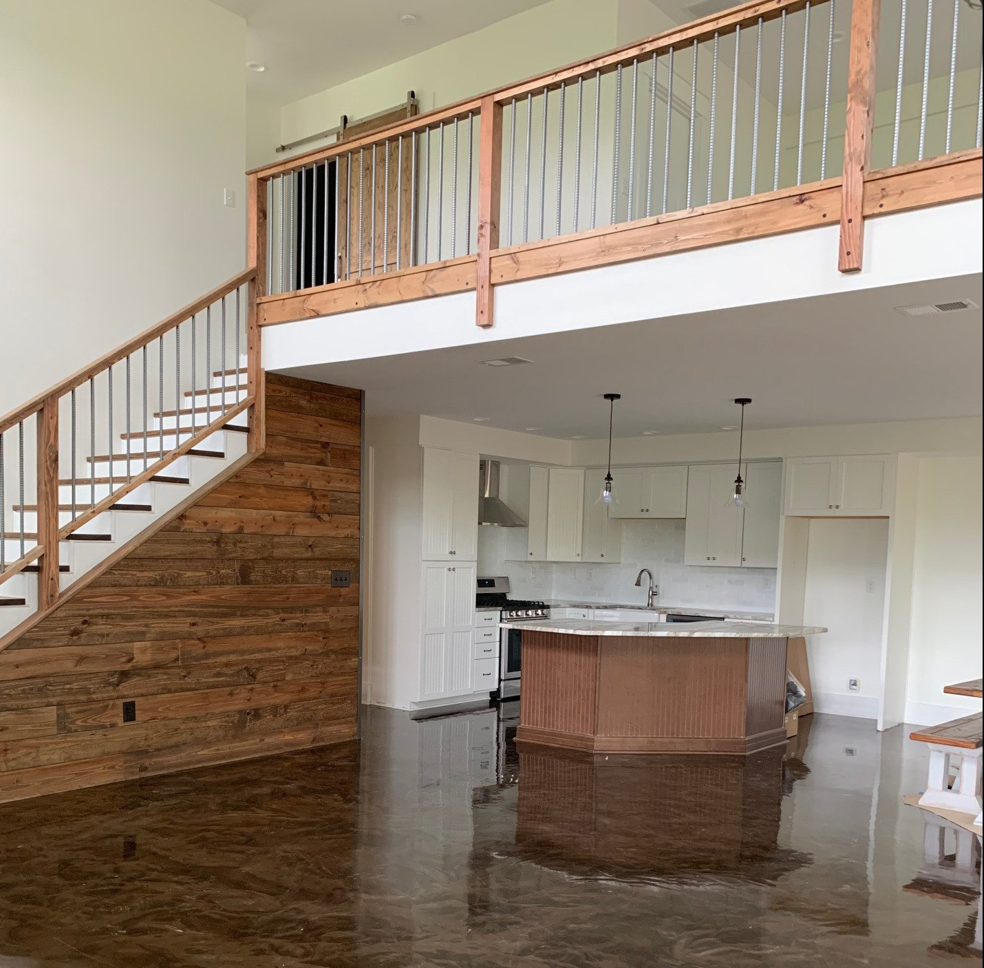 Interior shot of a modern home with a loft. Features wooden accents, a staircase, a kitchen island, and a polished concrete floor.