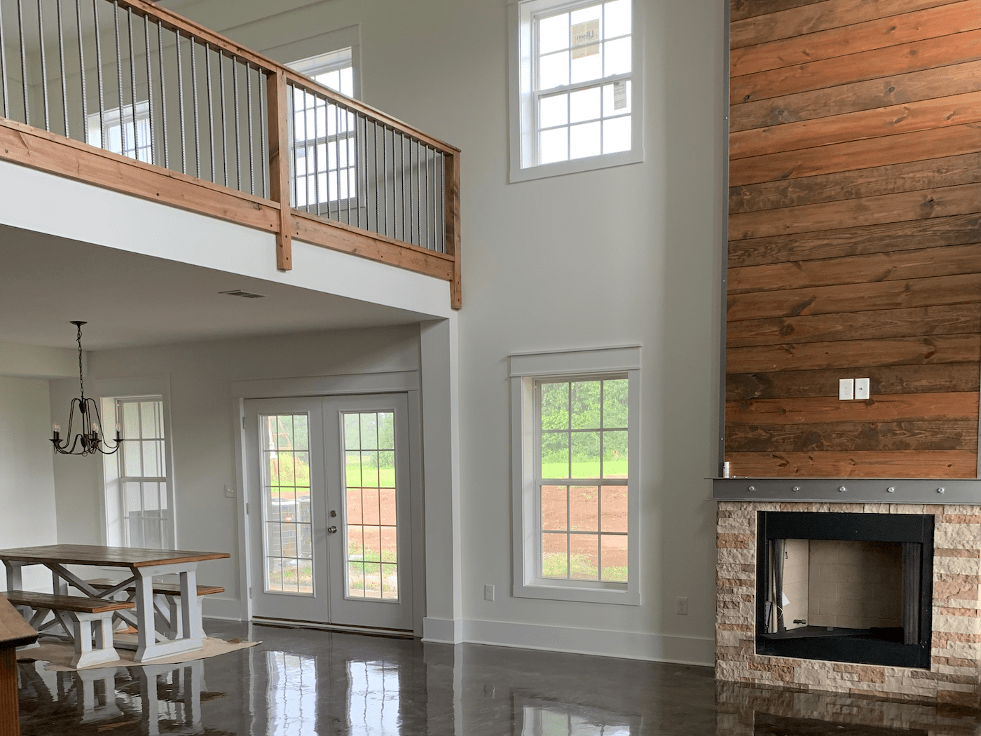 Bright interior with a high ceiling, featuring a loft, fireplace, dining area, and windows overlooking a green landscape.