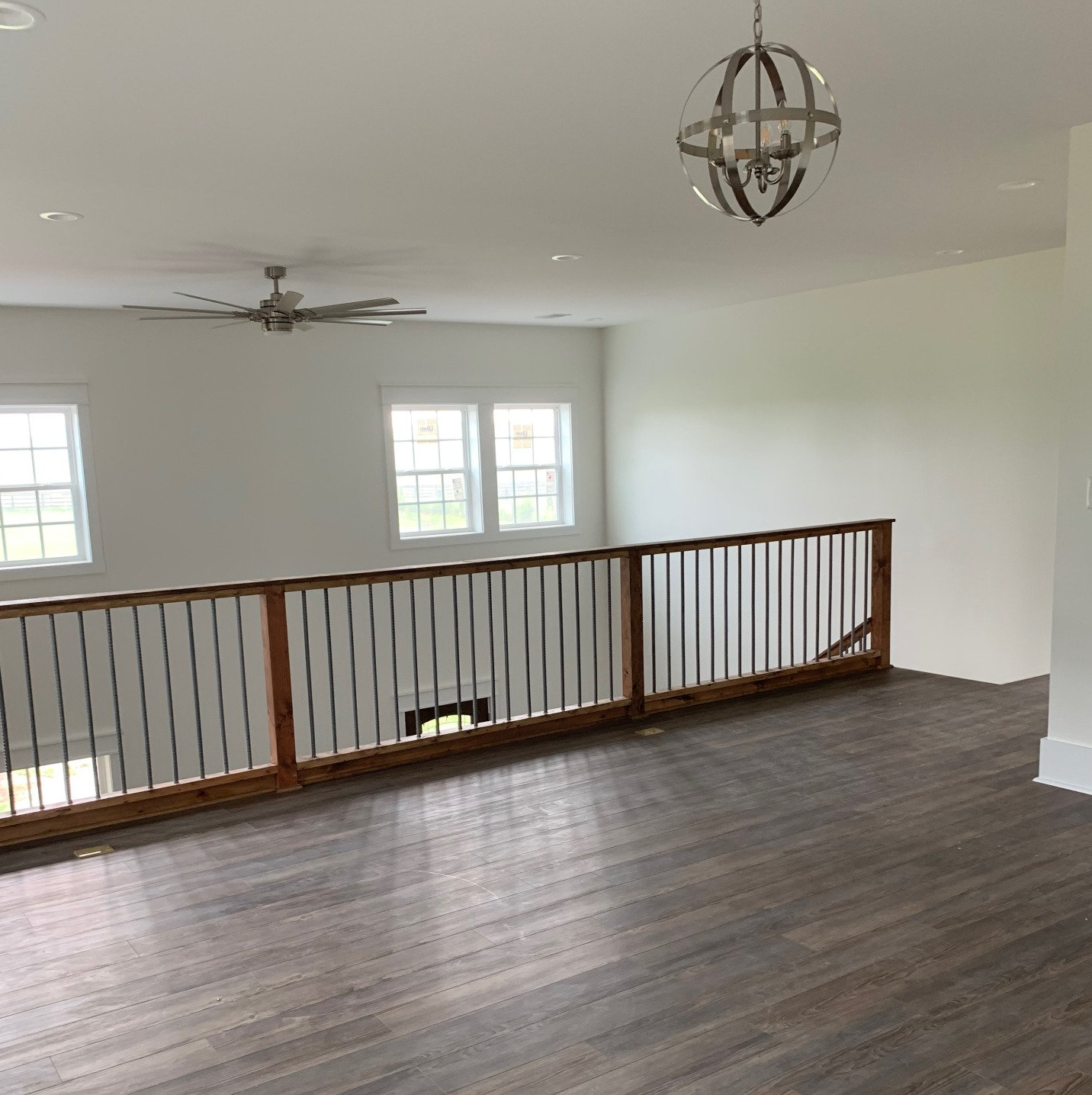 A modern, open-concept loft area with dark wood floors, a wooden railing, and white walls. A ceiling fan and spherical chandelier are visible.