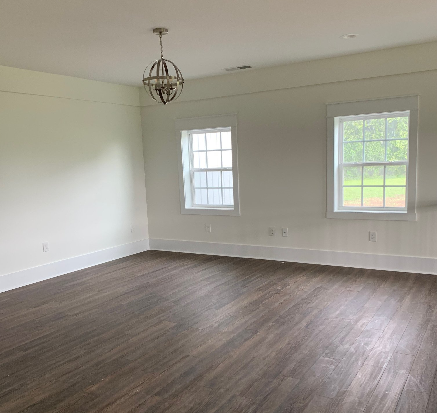 Empty room with hardwood floors, white walls, two windows, and a decorative chandelier.