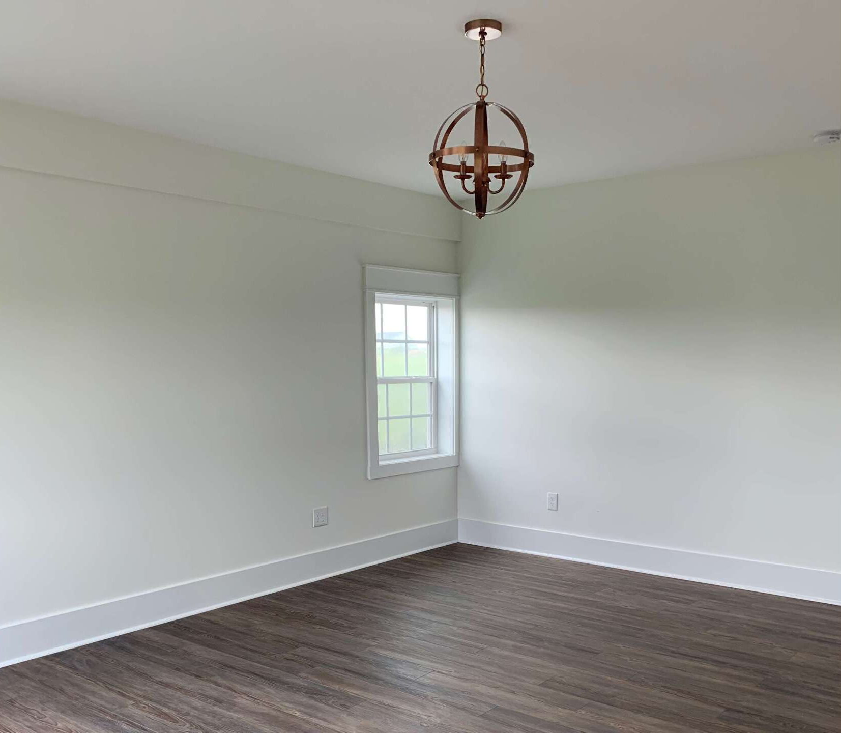 Empty room with white walls, dark wood floors, and a globe chandelier. A small window is on the left side.