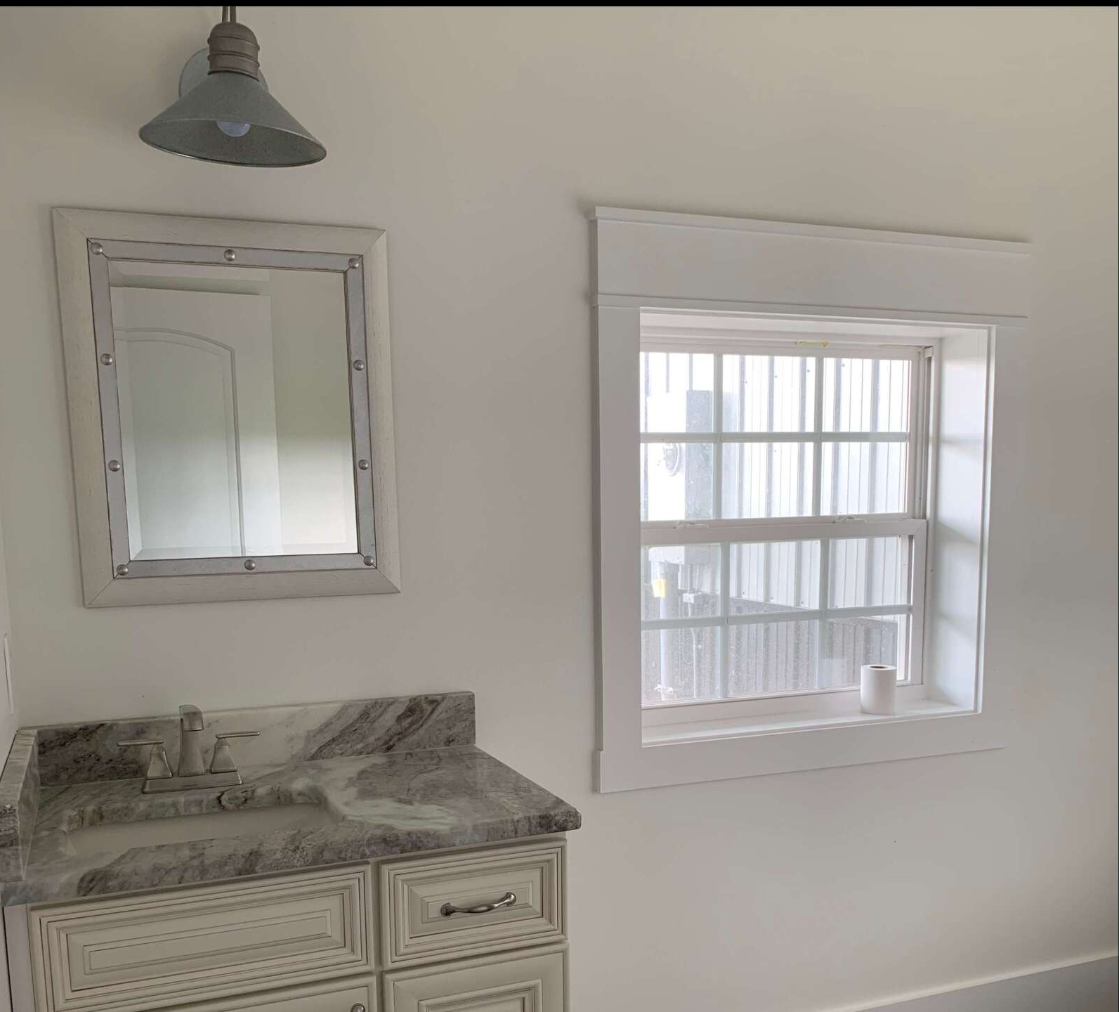 A small bathroom with a white vanity, window, and mirror. The countertop is light gray, and a metal light hangs above the mirror.