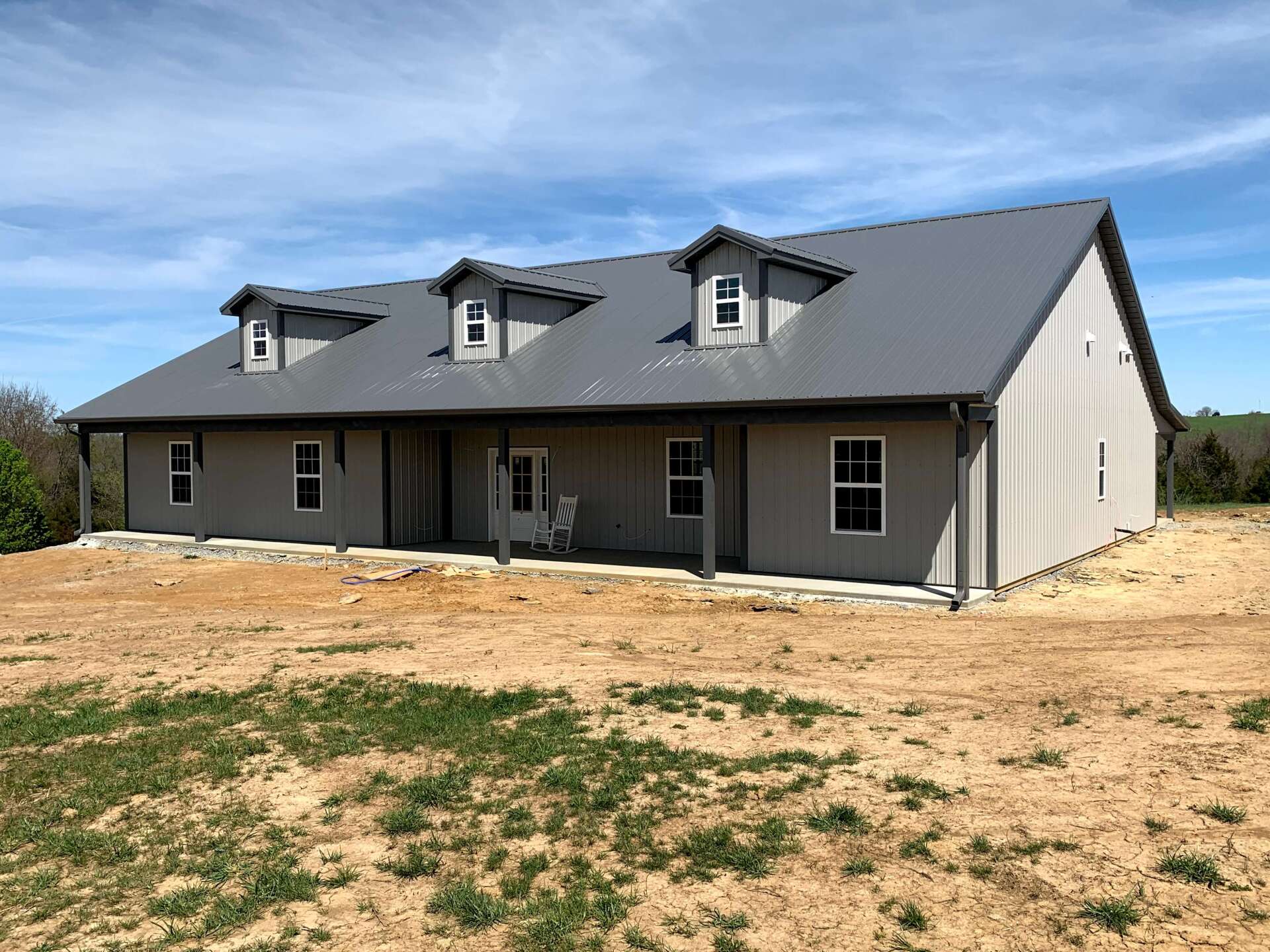 A long, gray, metal-roofed house with three dormer windows. It has a porch and sits on a dirt lot under a blue sky.