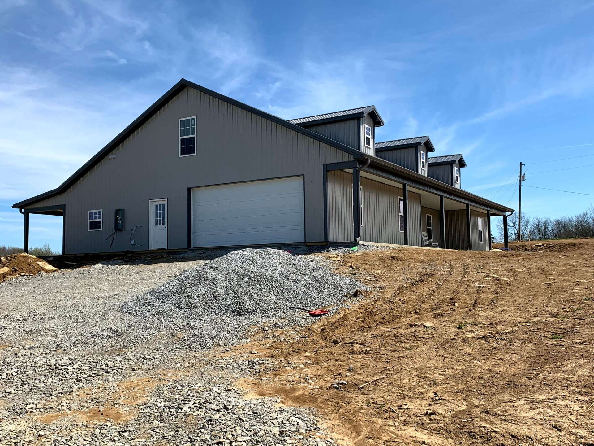 Gray metal building with a large garage door and a covered porch, under a blue sky. A gravel pile and dirt mound are in the foreground.