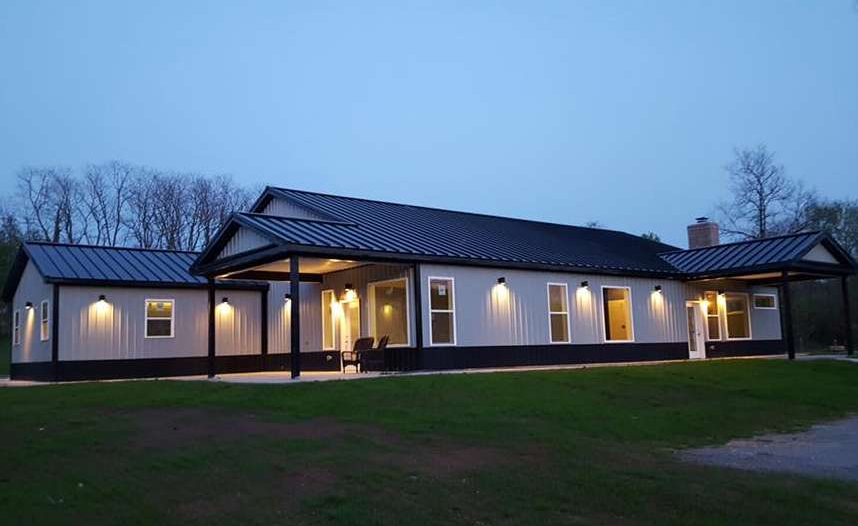 A modern, light gray house with a black roof and trim at dusk. Exterior lights are on, illuminating the building.
