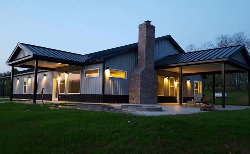 Modern house at dusk with metal siding and a brick chimney. The house has covered porches and warm interior lighting.