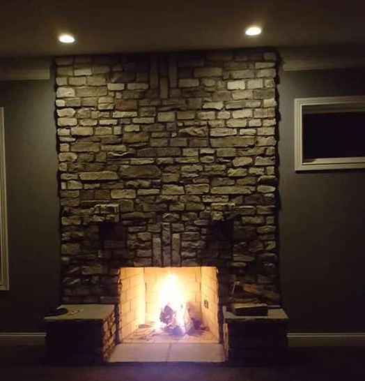 Stone fireplace with a burning fire, set in a dark room. Recessed lighting illuminates the stone and fire.