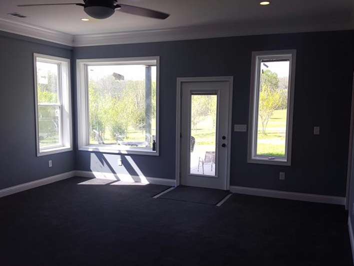 Interior view of a room with blue-gray walls, windows, a door, and dark carpet. Sunlight streams in, illuminating the space.