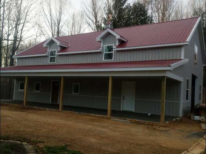 Two-story gray house with a red metal roof, dormers, and a covered porch with wooden beams. Set in a wooded area.