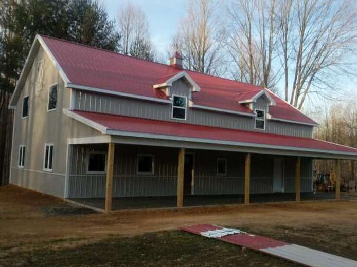 Two-story gray barn with a red roof and porch, standing in a grassy area with trees in the background.