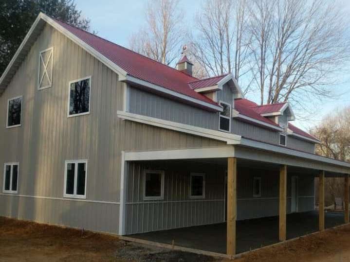 Two-story gray barn with red roof and white trim, featuring a porch, windows, and a cupola, set against a lightly wooded backdrop.