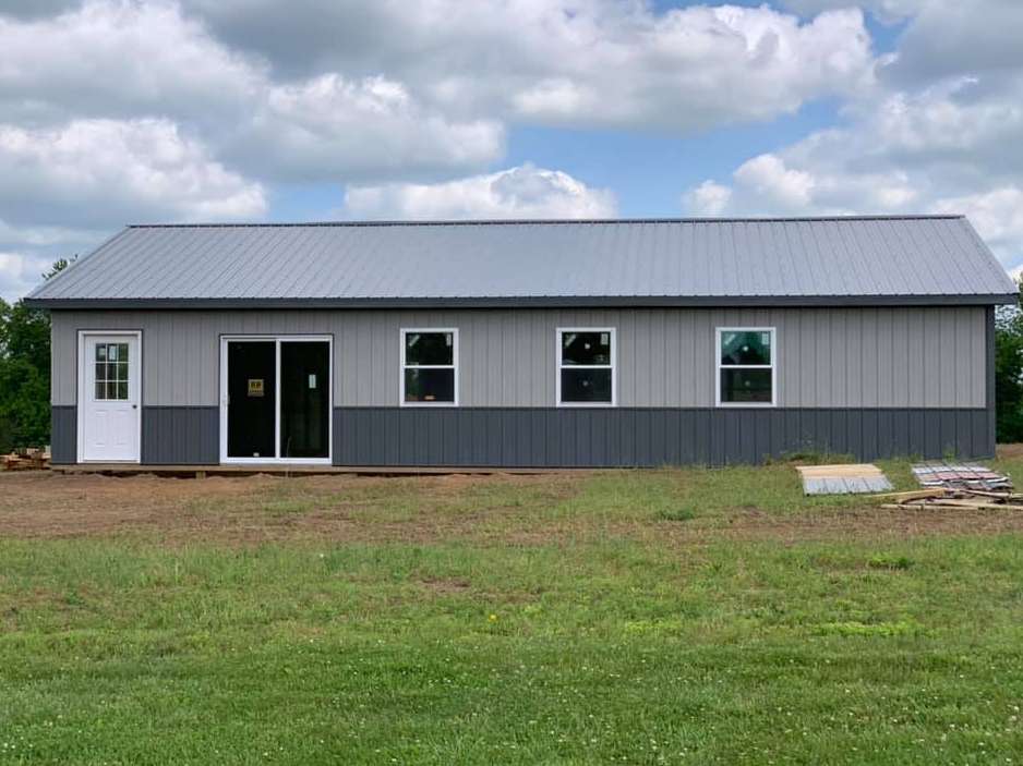 A gray and dark gray metal building with white doors and windows, set in a grassy field under a cloudy sky.