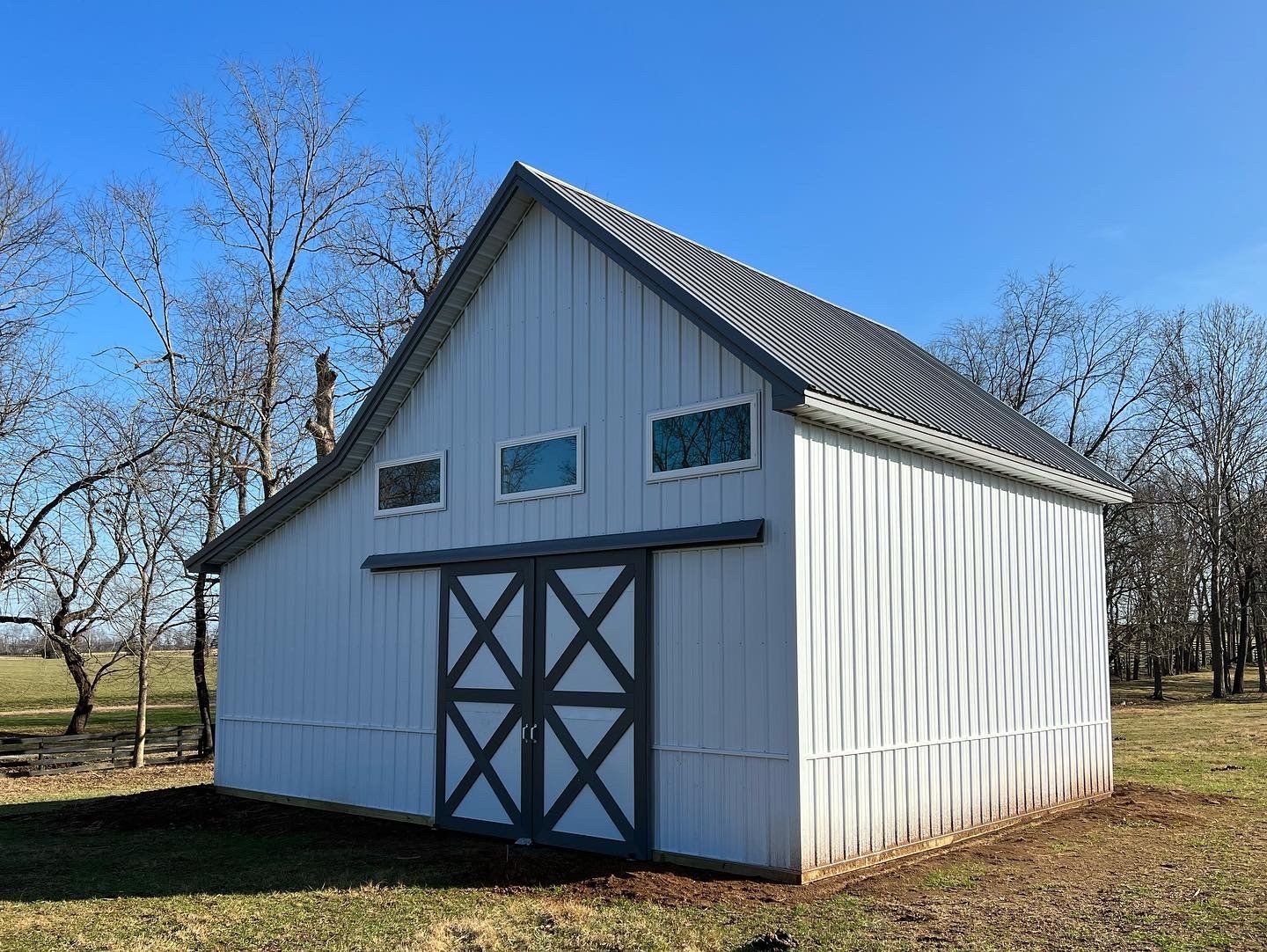 White barn with gray trim and doors, three small windows, set in a field on a sunny day.