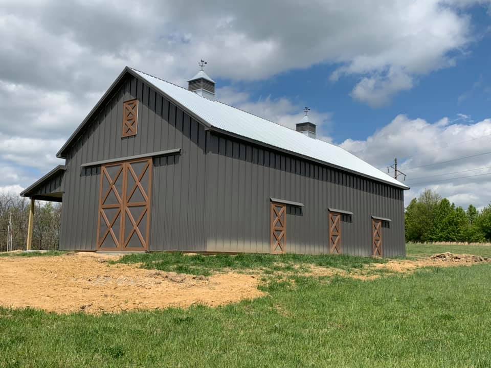 A gray barn with a silver roof and brown doors and trim stands on a grassy field under a cloudy sky.